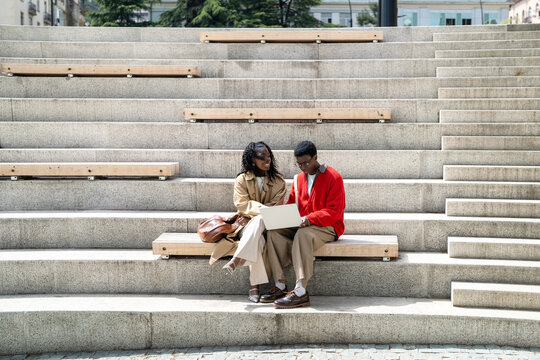Positive smiling african american students friends man and woman on daily break relaxing outdoors, black male nerd student and stylish black female discussing studies while working on laptop in campus