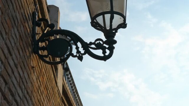 Wrought iron streetlight hangs from the corner of a brick building, with a clear blue sky and soft clouds providing a picturesque backdrop, highlighting urban architecture and design