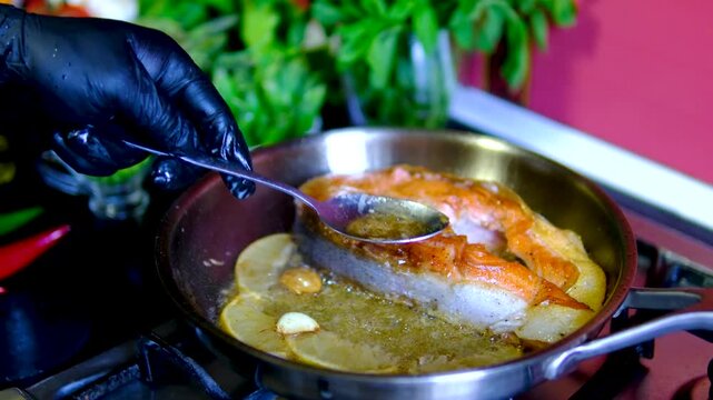 Salmon steak basted with butter by chef using spoon while garlic and lemon cook in hot pan during seafood recipe close up