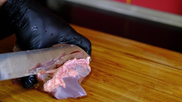 Chef in black gloves trimming connective tissue from fresh raw beef liver on wooden cutting board, cleaning organ meat before cooking, professional kitchen food preparation