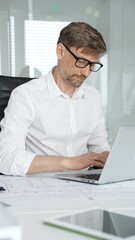 Obraz premium Professional architect or engineer working on a design project at an office desk, focused on his laptop with blueprints and a digital tablet visible