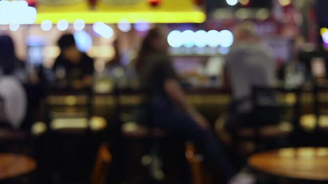 Blurred background of a busy bar or pub with customers sitting on stools at the counter. Soft bokeh lights and out of focus silhouettes of people enjoying nightlife, dining and drinking.