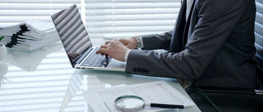 Business professional hands typing on a laptop keyboard. Working on financial documents. With a magnifying glass nearby. Representing analysis. Auditing