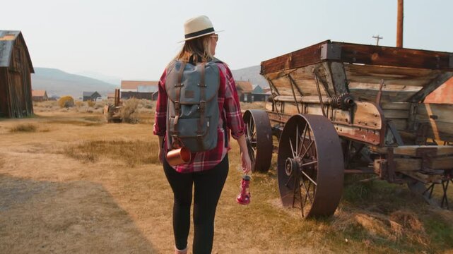 Young woman traveler with backpack and lantern walks through historic abandoned ghost town past weathered wooden wagon with rusty iron wheels.