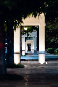Night walkway with glowing white arches forming a long symmetrical tunnel framed by greenery