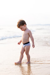 Young child standing on sandy beach, looking down at gentle waves along calm shoreline © Mikhail Mishchenko