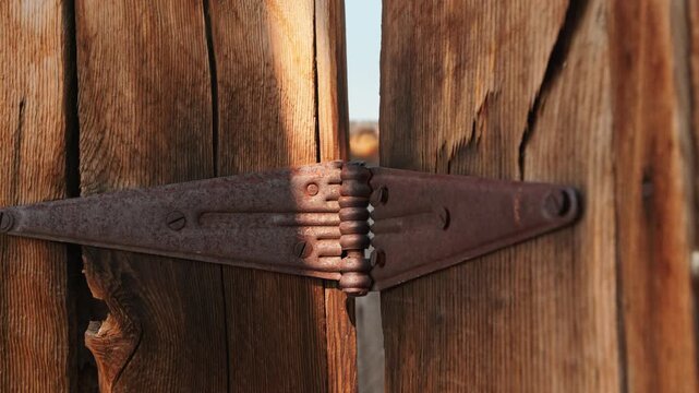 Old abandoned ghost town in America featuring close-up of rusty metal hinge on weathered wooden planks. Vintage iron hardware with patina shows age and decay in warm sunlight.