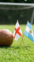 Fototapeta premium Vintage-style brown leather soccer ball next to small flags of England and Argentina on the grass.