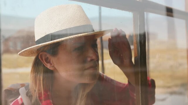 Girl walks through old ghost town in America peering through dusty window of abandoned building. Woman in straw hat and plaid shirt touches weathered glass pane in rural desert setting.