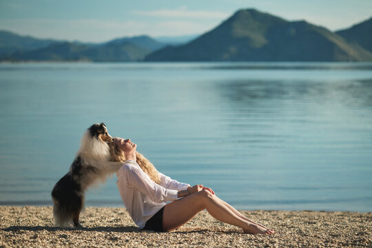 A woman enjoys a peaceful moment with her Shetland Sheepdog beside a scenic mountain lake. Relaxing outdoor lifestyle image featuring companionship and beautiful natural surroundings.