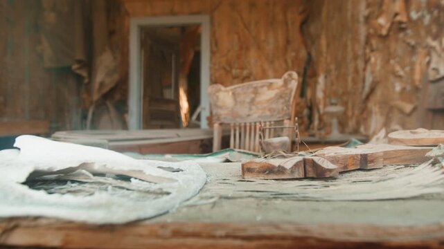 Old abandoned ghost town in America featuring decaying interior with peeling wallpaper and weathered wooden chair. Dusty table surface shows debris and aged fabric in deteriorating historic building.