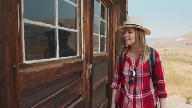 Girl walks through old abandoned ghost town in America examining weathered wooden structures. Tourist wearing straw hat and plaid shirt explores historic ruins in desert landscape on sunny day.