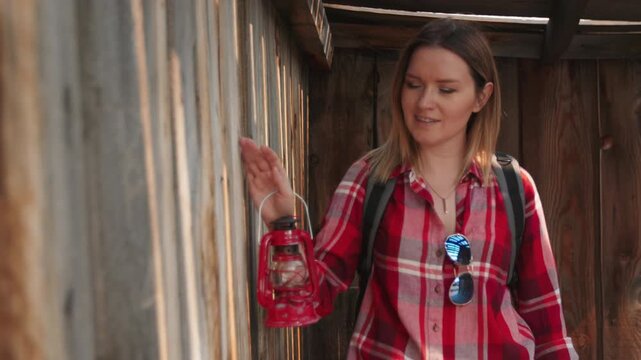 Young woman in red plaid shirt holding vintage lantern explores rustic weathered wooden corridor of abandoned ghost town, touching textured gray planks while warm golden sunlight filters through gaps.