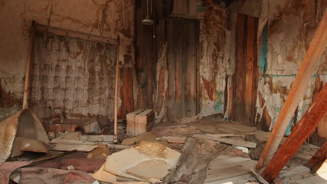 Old abandoned ghost town in America featuring decaying room interior with rusty metal bed frame, peeling wallpaper, scattered debris on wooden floor and hanging vintage light fixture.