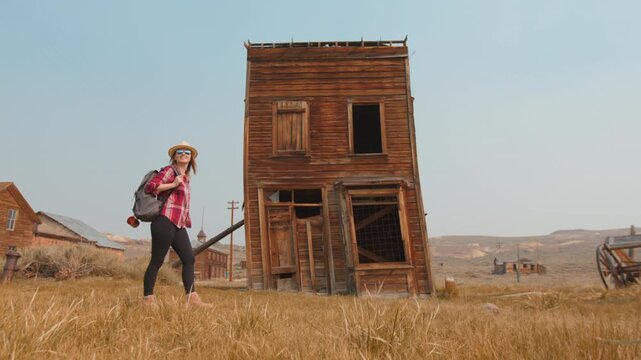 Young woman traveler with backpack and straw hat walks through golden dry grass past weathered two-story wooden building in historic abandoned ghost town under hazy blue sky during golden hour.