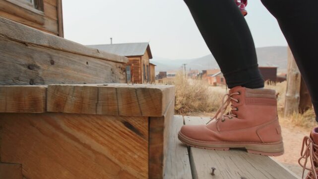 Girl walks through old abandoned ghost town in America. Pink boots step on weathered wooden porch of historic western building with rustic structures and mountains in background on sunny day.