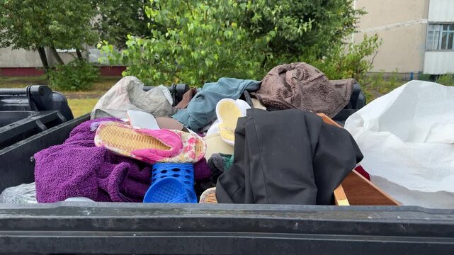 Overflowing black dumpster filled with discarded clothes, slippers, and towels in a residential yard. Waste disposal, trash, poverty, decluttering, social issue