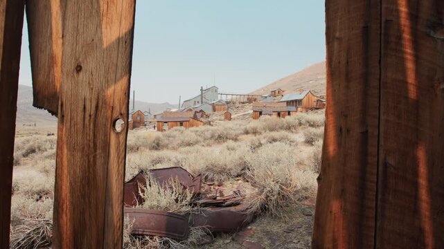 Old abandoned ghost town in America viewed through weathered wooden posts. Rustic mining settlement with decaying buildings and rusty metal debris scattered among sagebrush in arid desert landscape.