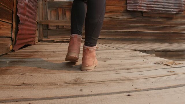 Girl walks through old abandoned ghost town in America wearing pink boots on weathered wooden boardwalk. Rustic wooden buildings and corrugated metal siding create authentic western atmosphere.