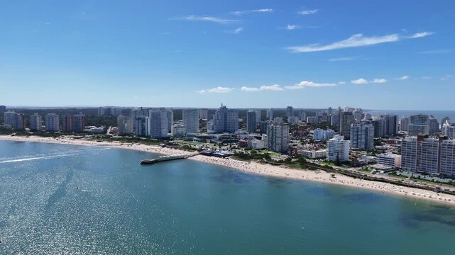Drone view of Mansa Beach, the iconic wooden pier, turquoise waters and the modern skyline of Punta del Este on a sunny summer day in Uruguay.