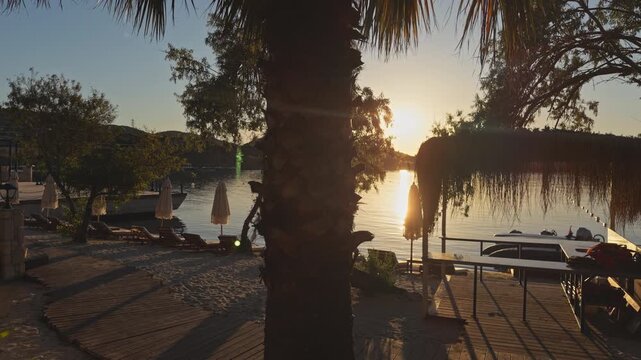 Bright golden sun flares behind a tall palm tree trunk and fronds at a tropical beach resort, reflecting warmly across calm water beside a thatched cabana and sandy loungers, creating a dramatic intim