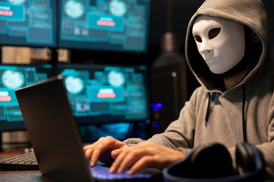 Masked hacker attempts a data breach in a tech environment, anonymously writing malware code on a laptop and targeting sensitive digital information.