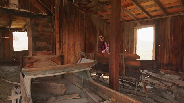 Young woman wearing straw hat and plaid shirt explores weathered rustic wooden barn interior with exposed beams and scattered debris in abandoned ghost town bathed in warm natural daylight.