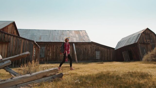 Young woman in straw hat and red plaid shirt explores abandoned ghost town with rustic wooden barns and weathered buildings under hazy blue sky, golden dry grass covering the landscape at sunset.