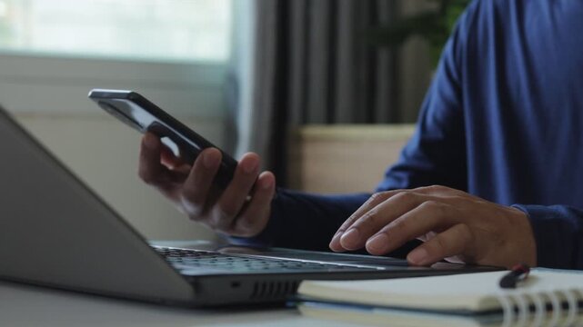 Closeup of businessman using smartphone while typing on laptop in modern workspace, concept of multitasking, remote work, digital communication, productivity, cybersecurity and technology lifestyle.