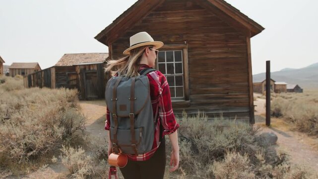 Girl walks through old abandoned ghost town in America exploring weathered wooden structures. Female traveler with backpack and hat touches rustic building walls amid desert sagebrush landscape.