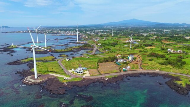 Aerial forward flight along winding Sinchang coastal road with wind turbines green farmland car driving and Hallasan mountain on Jeju Island volcanic shore in summer