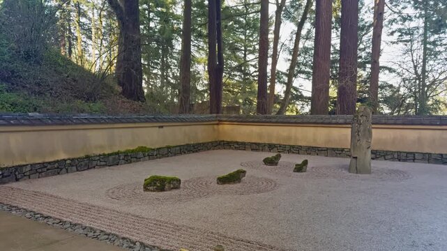 Raked gravel zen garden with mossy stones enclosed by walls beneath towering forest trees, establishing pan right