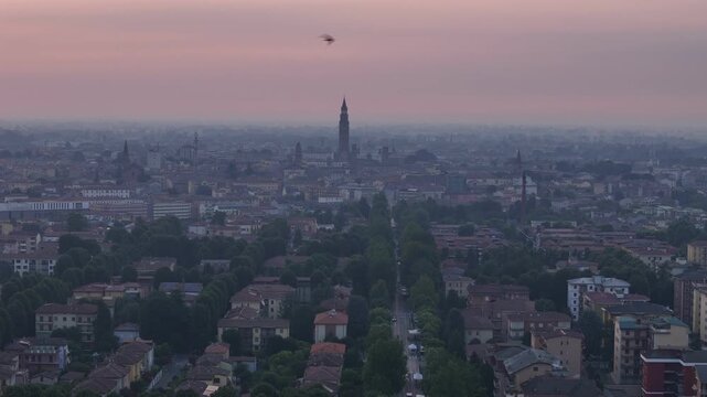 Drone pulls back above the historic skyline of Cremona, Lombardy, Italy, revealing the Torrazzo, Cremona Cathedral, Viale Po and hazy sunrise conditions during the 2026 summer heatwave, birds crossing