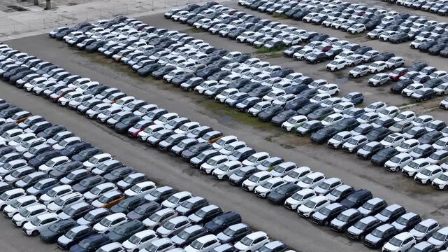 Oblique aerial view of a large automobile storage yard with thousands of parked passenger vehicles arranged in organized rows for fleet logistics and distribution, drone oblique flyover.