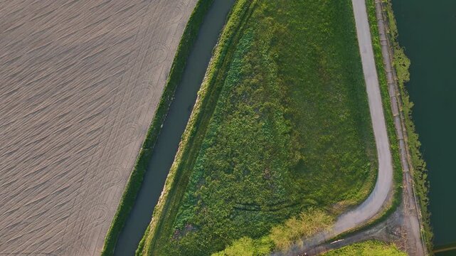 Top-down drone view of an irrigation canal crossing Po Valley farmland in northern Italy, bordered by cultivated fields, grassy levees and a rural service road, static overhead shot.