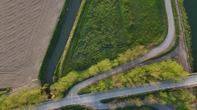 Drone ascends vertically above an irrigation canal, cultivated farmland, grassy levees and intersecting rural roads in the Po Valley, northern Italy, revealing geometric agricultural patterns.