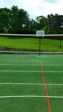 An empty outdoor multisport court featuring a volleyball net and basketball hoop, surrounded by green grass and trees.