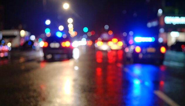 Blurred city traffic lights at night with glowing red and blue police vehicle sirens on a wet rainy urban street