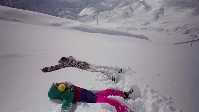 Two people dressed in colorful winter clothes fall backward and lie in fresh snow on a bright day in the snowy Mendoza mountains.