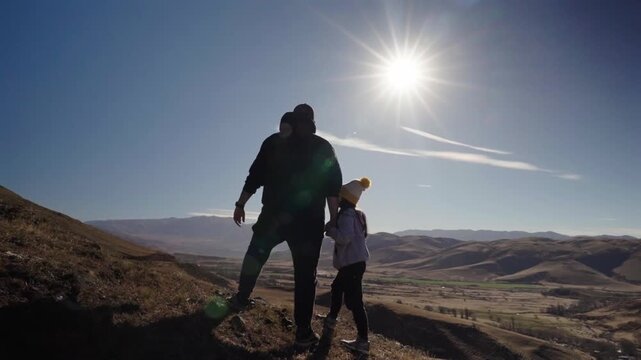 A cinematic silhouette of a father and his young daughter holding hands while looking at a vast valley during golden hour. Authentic family wellness and outdoor adventure.