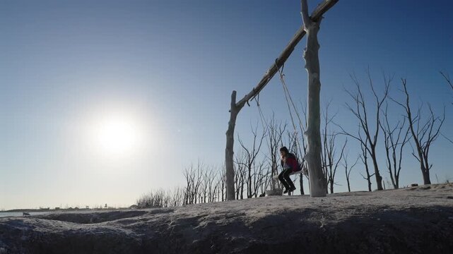 A young child plays on a rustic wooden swing set in a barren, cinematic landscape with dead trees under a bright golden hour sun. Ideal for themes of solitude, climate change, and childhood.