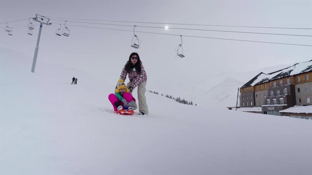 Parent helps child on sled at snowy ski resort beneath chairlifts, winter, pushes her down hill, slow motion
