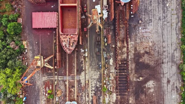 Large boats are under construction at the shipbuilding site with materials scattered around. Workers are moving tools while machines are parked near the workshop.