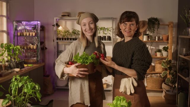 Medium slowmo portrait of Caucasian young woman and her mother holding fresh vegetables in indoor greenhouse, smiling at camera. Video is ideal for promoting sustainable urban farming