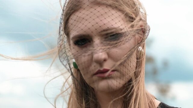Young woman in black net veil stands outdoors with downward gaze while breeze lifts hair and sheer mesh across face, creating diamond shadows, soft highlights, muted mood, refined fashion feel