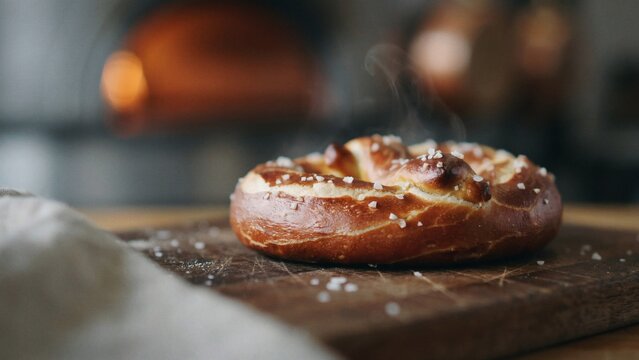 A close-up photo of a salted pretzel on a wooden cutting board, with a blurred bakery background.
