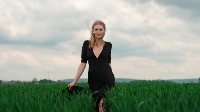 Young woman moves along narrow path through tall vibrant grass in wide rural field, alternating walking and kneeling while adjusting birdcage veil, creating calm moody fashion scene