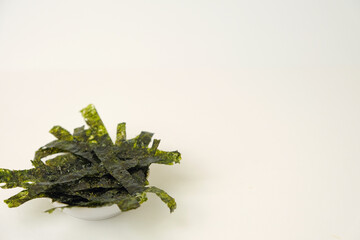 Dark green seaweed flakes rest in a small white bowl. Bright white background enhances the natural texture. Clean, minimalist composition ideal for health or food themes © Nurma