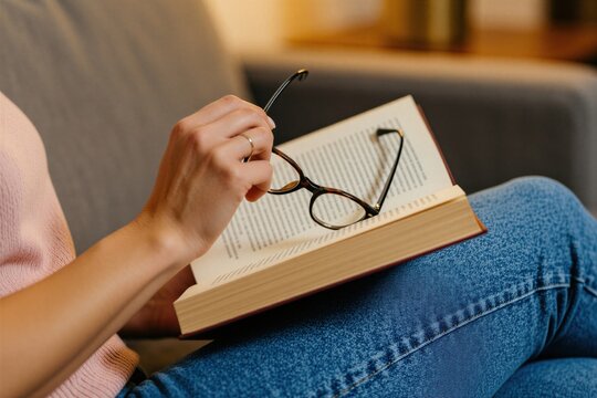 Young woman sits on soft sofa at home, holds open book. Female reader kepic glasses in hand relaxing on couch. Person spends leisure time studying literature indoors. Comfortable student enjoys quiet