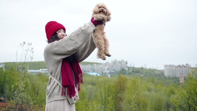 Happy woman dressed in a beige sweater, red knit hat and magenta scarf, stands on a mossy hilltop amid low heath vegetation, holds and shows the small terrier dog. Overlooking valley and buildings